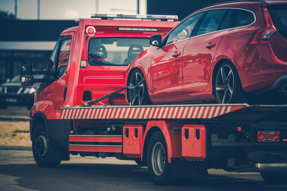 A red tow truck from a local towing company is transporting a red car on its flatbed. The scene is set outdoors, with a blurred background suggesting a roadside or parking area. The vehicles are facing left, highlighting the rear view of the tow truck, showcasing efficient towing services.