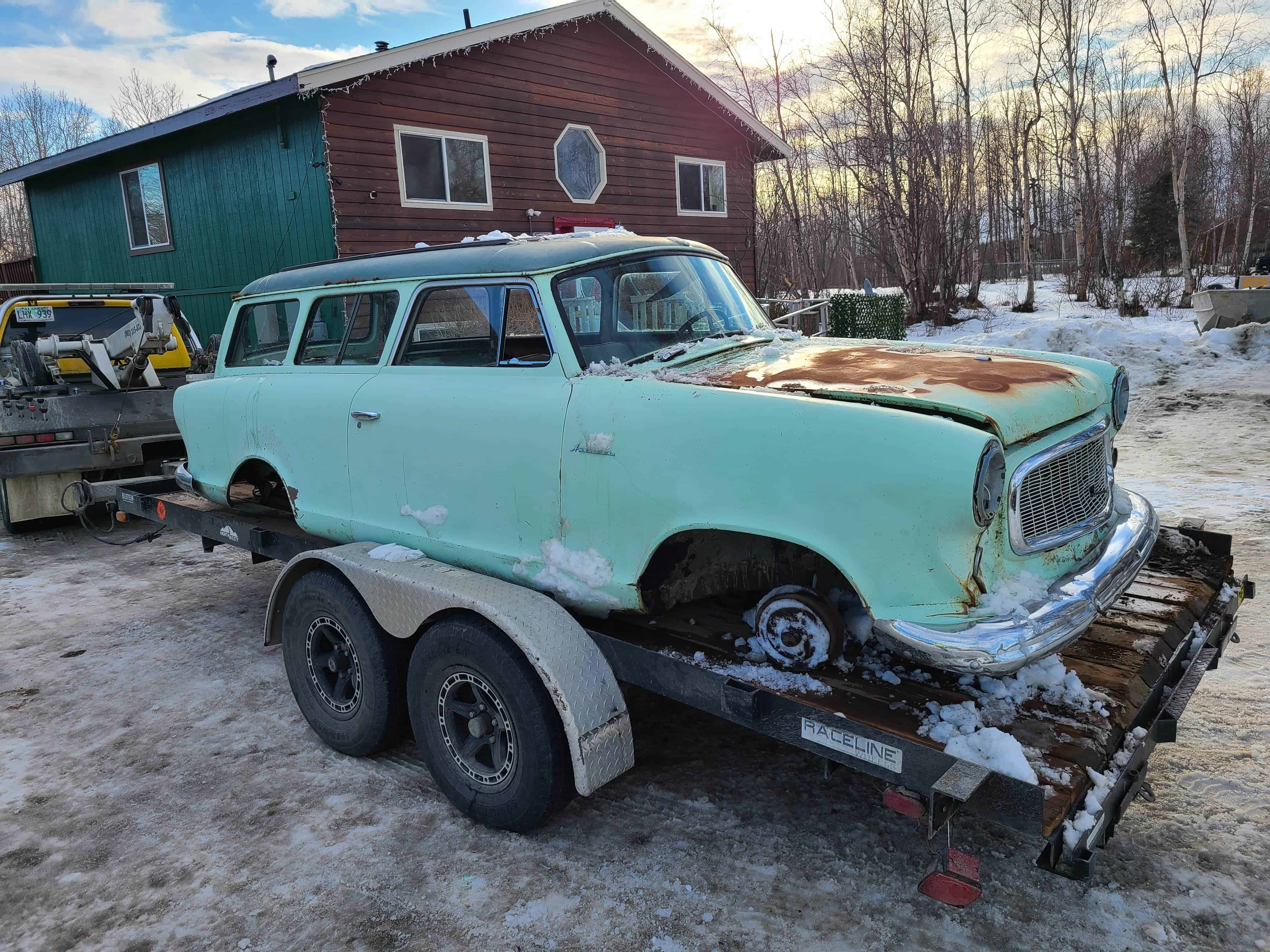 A vintage, mint green wagon with visible rust sits on a trailer surrounded by snow. The backdrop highlights a wooden house and trees on a cloudy day, showcasing the expertise of a local towing company in ensuring even the most cherished vehicles reach their destination safely.
