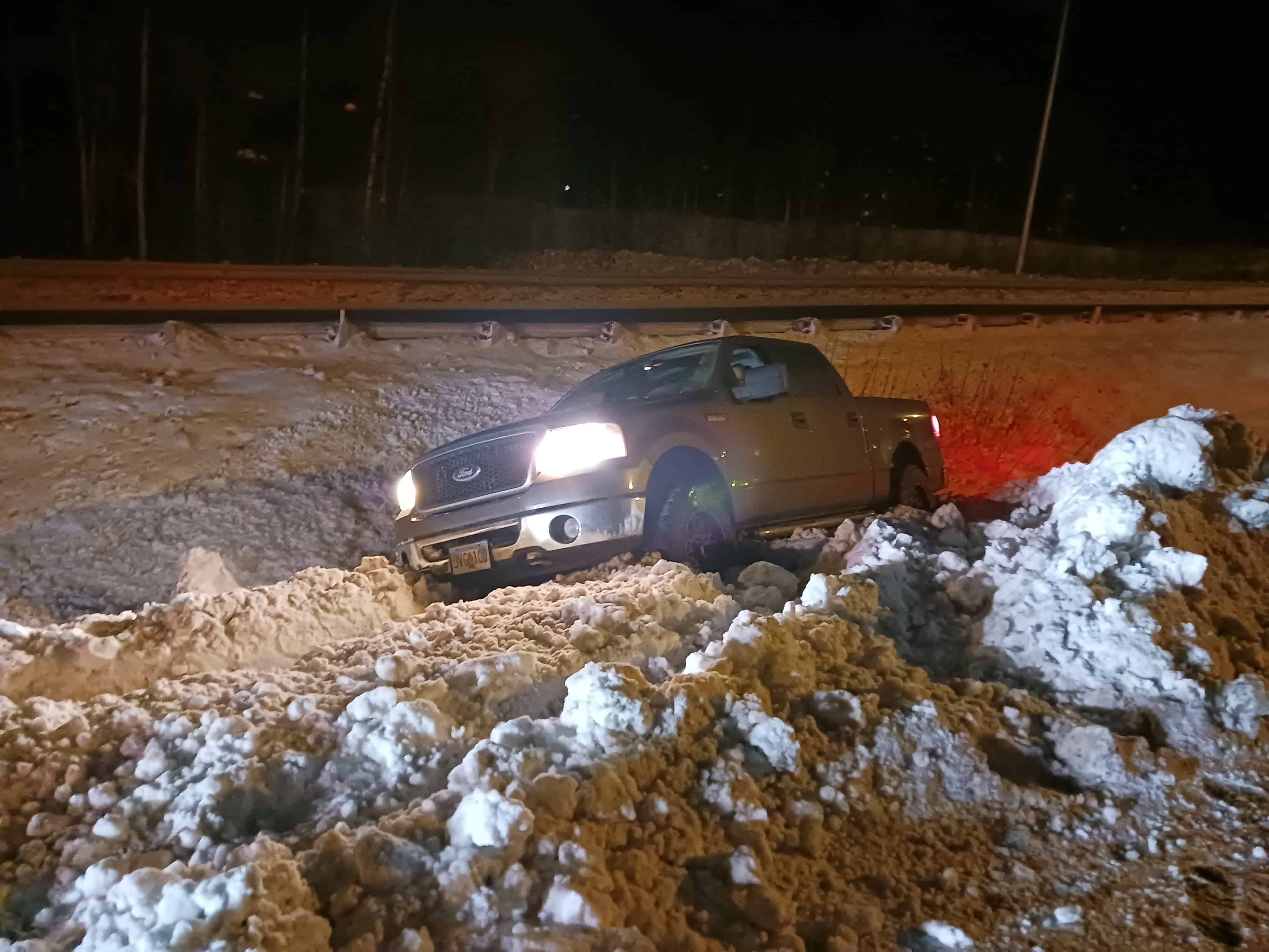 A pickup truck is stuck in a snowy ditch at night, its headlights illuminating the snow while red taillights glow. A metal guardrail and trees form a backdrop as an emergency roadside assistance crew arrives to provide towing services, cutting through the cold with timely expertise.