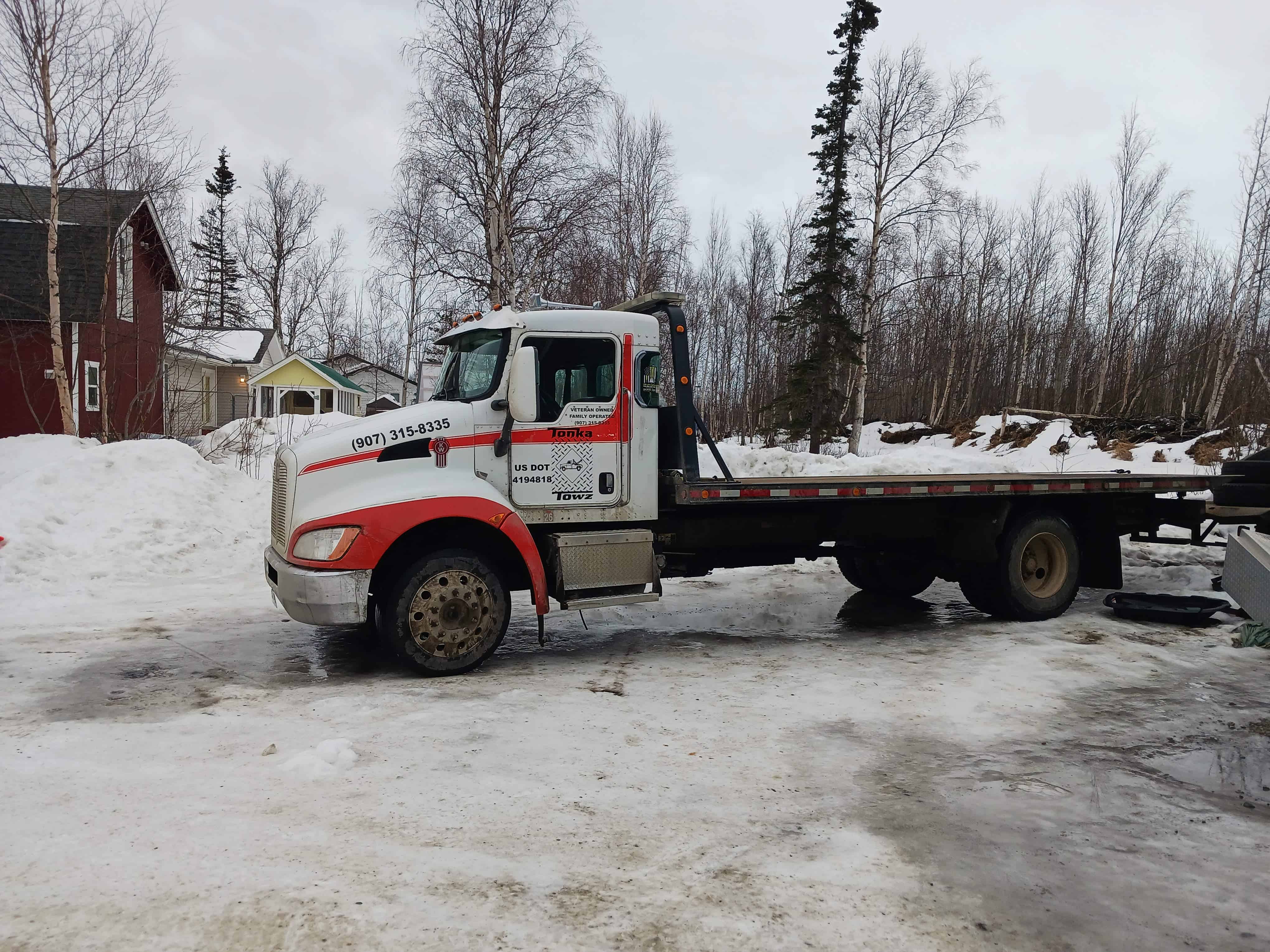 A red and white flatbed tow truck from a local towing company is parked on a snowy, icy driveway surrounded by leafless trees. Nearby, there is a red house and a smaller structure. The overcast sky enhances the wintry atmosphere, ideal for showcasing reliable emergency roadside assistance.