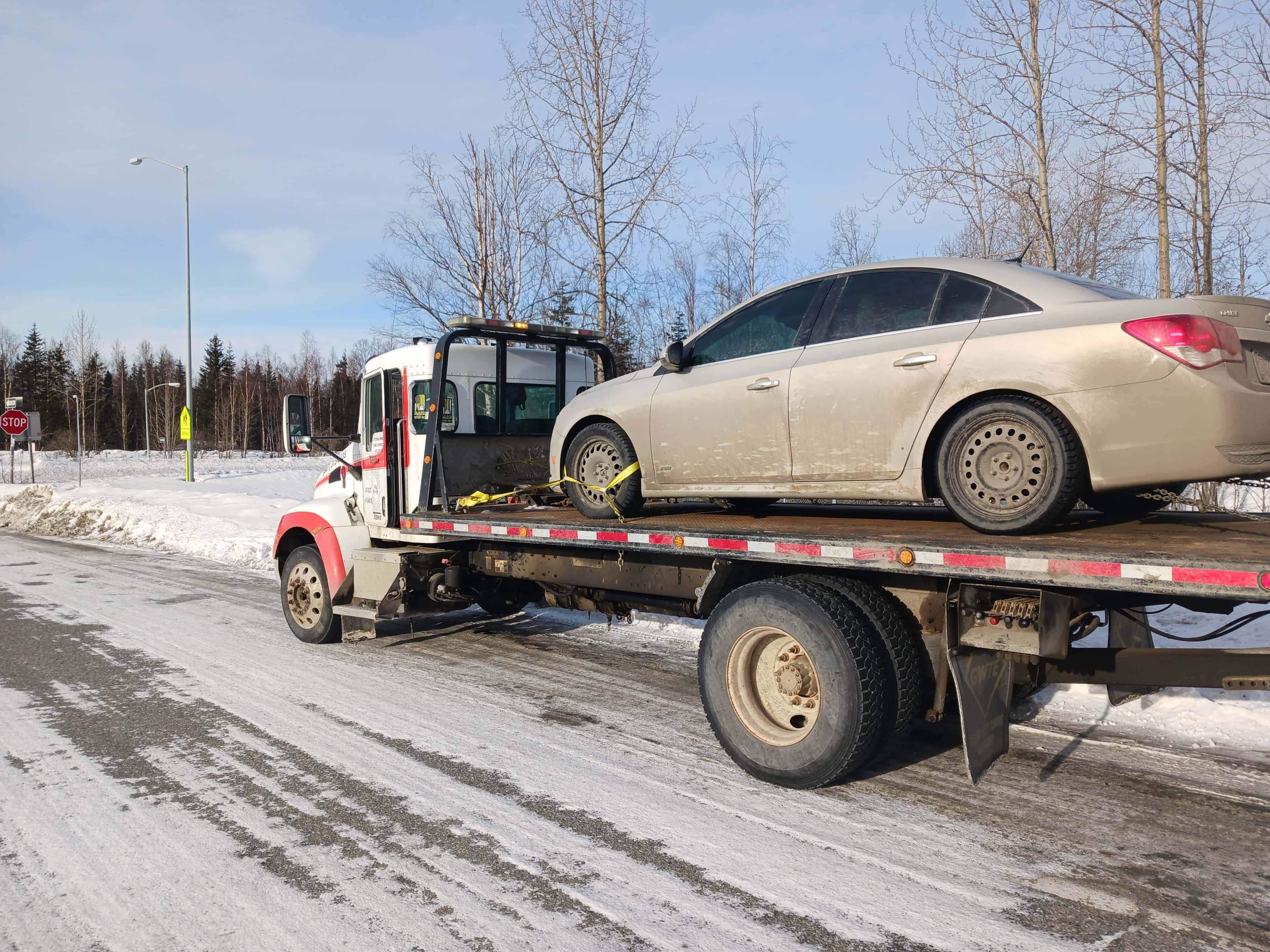 A towing company truck transports a dirty sedan on a snowy road. The sedan is secured on the flatbed, surrounded by snow-covered ground and bare trees under a clear blue sky, highlighting expert emergency roadside assistance in action.