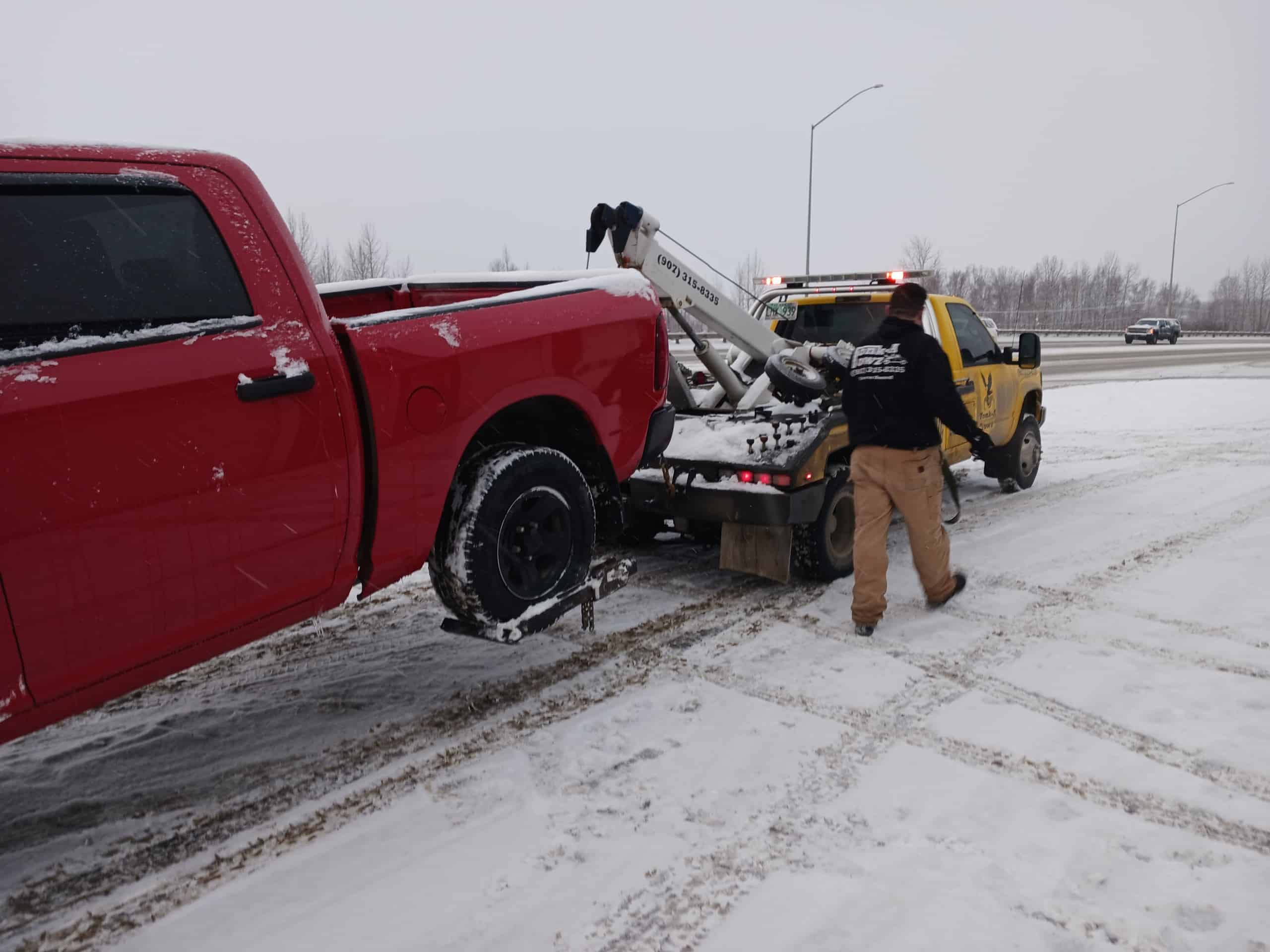 A yellow tow truck from a local towing company is towing a red pickup truck on a snowy road. A person in a jacket and boots is assisting with the emergency roadside assistance. The area is blanketed in snow, with bare trees visible in the background.