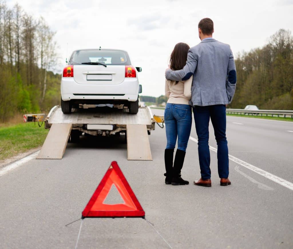 A couple stands on the roadside, watching their white car being loaded onto a tow truck. A red warning triangle marks the scene on the highway. Trees line the background as emergency roadside assistance efficiently manages the situation, ensuring safety and swift towing services.