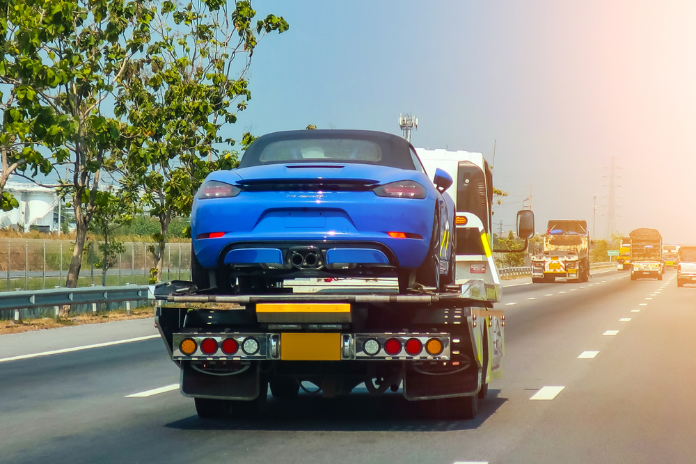 A blue convertible sports car is being transported by a towing company on a flatbed along a sunny highway. Trees line the road, and several vehicles are visible in the distance, exemplifying efficient emergency roadside assistance in action.