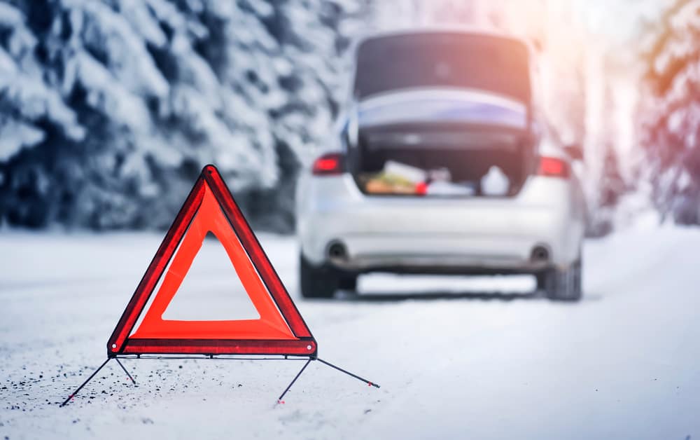 A warning triangle stands on a snowy road with a blurred car in the background, its trunk open. Snow-covered trees line the roadside under a soft, sunlit sky, as emergency roadside assistance arrives to aid those in need.