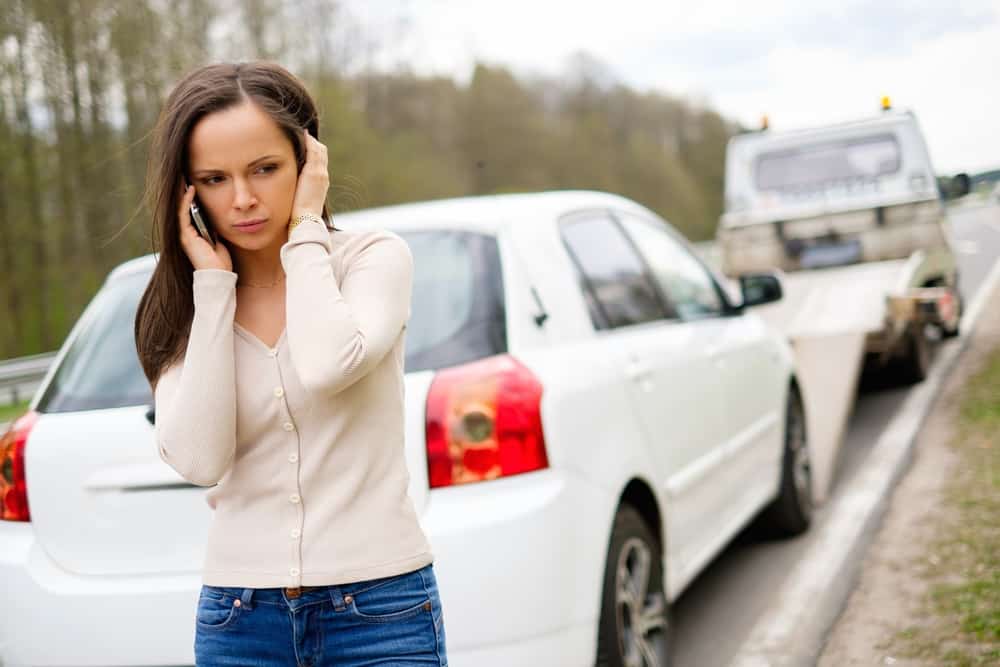 A woman stands by the roadside, looking concerned as she talks on her phone. A white car is parked behind her while a towing company truck is in the process of towing another vehicle. Trees line the background under a cloudy sky, illustrating the urgency of emergency roadside assistance.