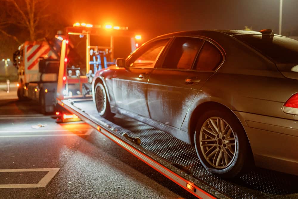 A dark-colored sedan is being loaded onto a flatbed tow truck at night, provided by a local towing company. The tow truck's bright orange lights illuminate the scene, reflecting off the car's surface. The dimly lit surroundings suggest a roadside or parking area in need of emergency roadside assistance.