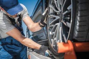 A mechanic in a blue cap, gloves, and overalls, representing a top-notch towing company, inspects a tire of a black car on a ramp. He adjusts the wheel with focused attention under the clear blue sky.