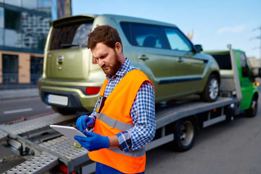 A man in an orange safety vest and blue gloves uses a tablet while standing next to a tow truck, providing emergency roadside assistance. The truck is transporting a green SUV. Urban buildings and a street are visible in the background, highlighting the towing company's efficiency.