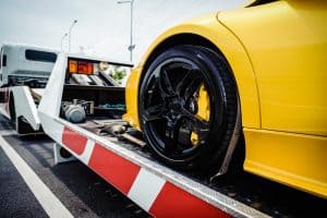A close-up of a sleek yellow sports car on a tow truck, showcasing its shiny black rear tire and part of the body. The truck's ramp, adorned with red and white stripes, hints at professional towing services. The background includes street signs and a cloudy sky.