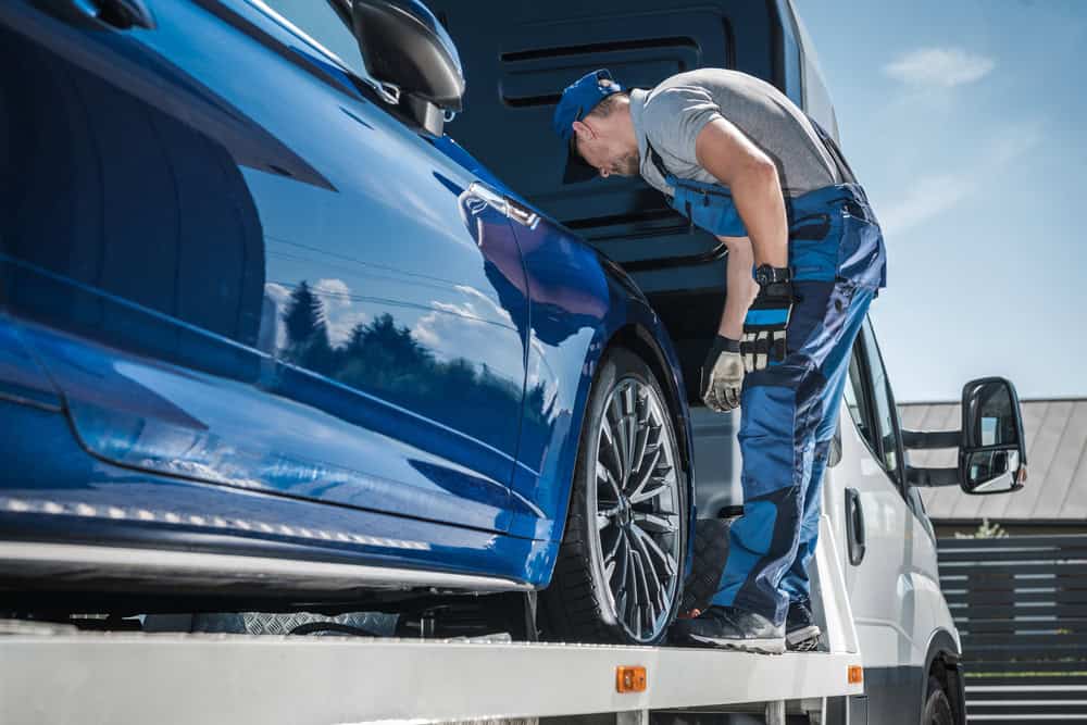 A mechanic wearing blue overalls and a cap inspects the wheel of a blue car on a flatbed truck, providing emergency roadside assistance. The scene is outdoors, with clear skies as a backdrop to the efficient towing services being expertly carried out.