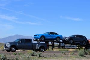 A gray truck from a reputable towing company is hauling a trailer with a blue convertible car and a dark SUV. They're traveling on a road with mountainous terrain in the background under a clear blue sky.