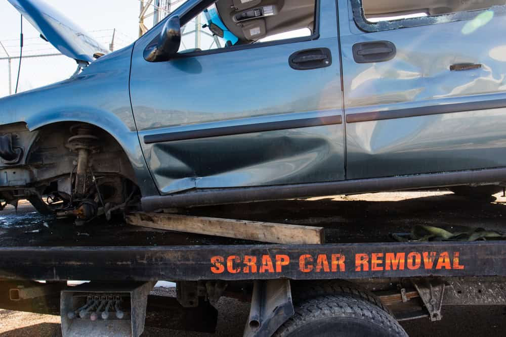 A damaged blue car is loaded onto a flatbed truck from a towing company labeled "Scrap Car Removal." Missing a front wheel with its hood open, exposing the engine, the scene suggests the vehicle is being transported for scrapping.