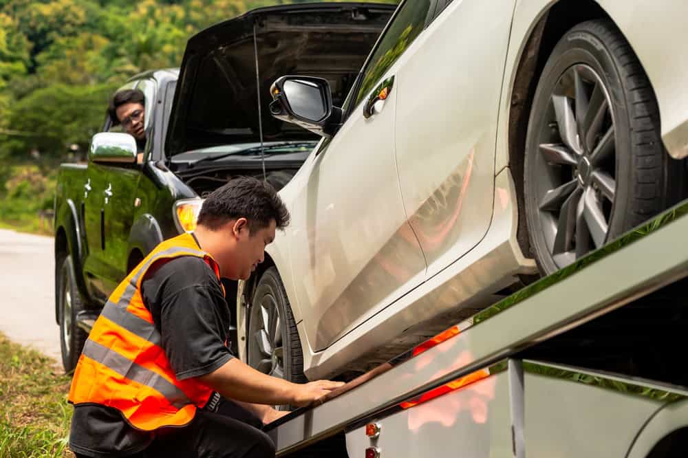 A man in a reflective vest operates the controls of a tow truck, loading a white car as part of an emergency roadside assistance task. Another person in a black vehicle observes the process, showcasing the efficiency of this towing company's services against a backdrop of roadside greenery.