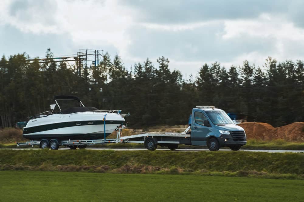 A blue van from a towing company is transporting a large white and black boat on a flatbed trailer along the road. Trees and a construction site with mounds of dirt are visible in the background under a cloudy sky.