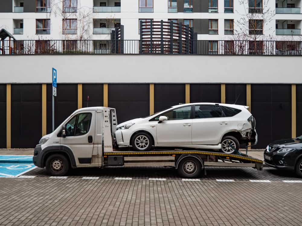 A white car with rear damage is loaded on a tow truck from a local towing company, parked on the street. Modern architecture and balconies create the backdrop, highlighting efficient towing services amidst urban life.