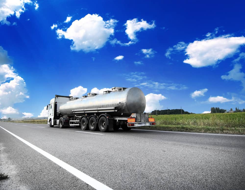 A shiny, silver tanker truck drives on a rural highway under a bright blue sky with scattered white clouds. Green fields and trees line the road, creating a serene landscape, as an emergency roadside assistance vehicle follows closely behind, ready to offer support if needed.