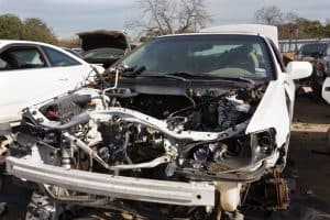 A dismantled white car, with its front parts removed to reveal the engine bay, is parked outdoors in what appears to be a scrapyard or repair area. The scene, surrounded by other vehicles and trees, hints at the expertise of a reliable towing company nearby.